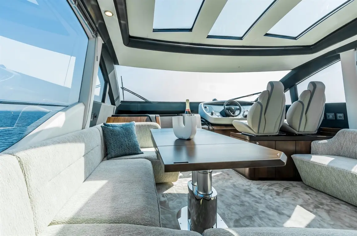Luxurious boat interior with white sofas, a coffee table, a champagne bottle, and a modern helm with a panoramic view.